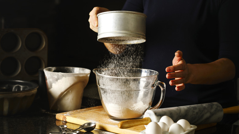 A baker sifts flour into a bowl on a kitchen cutting board