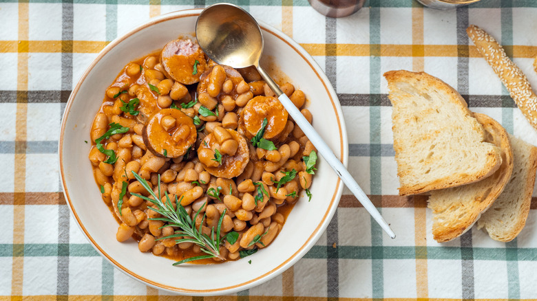 Bowl of beans with slices of sausage and herbs on top with bread on the side