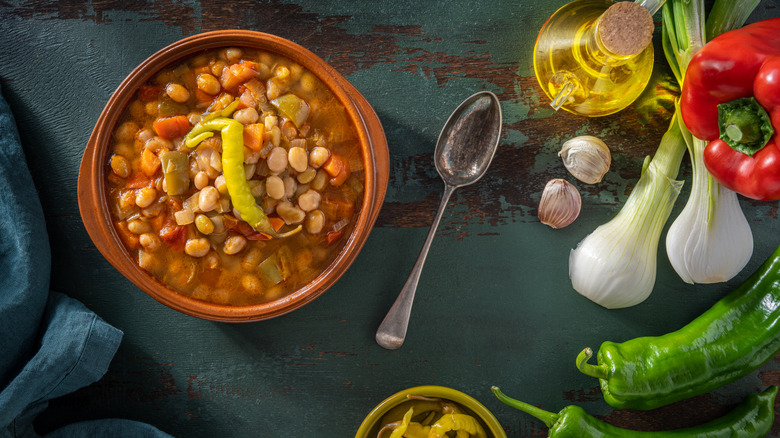 A bowl of beans along with some garlic cloves, oil, peppers, and onions