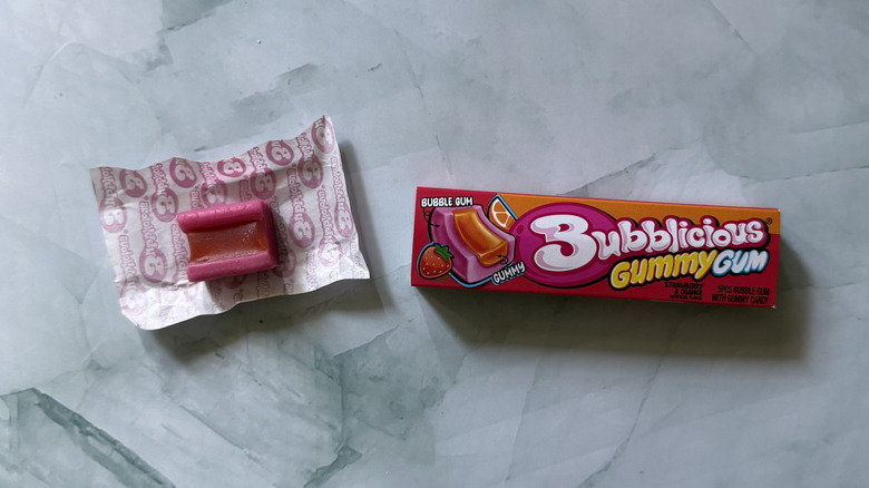 Pack of Bubblicious Gummy Gum in Strawberry & Orange flavor next to an unwrapped piece of the gum on countertop