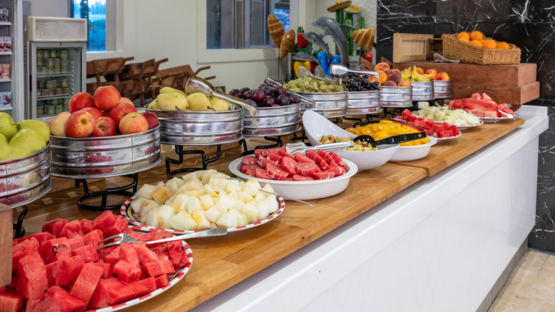 Several fruit platters sit on a buffet table, many with a set of tongs inside for serving.