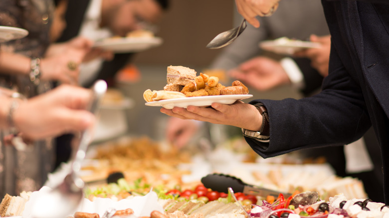 A man holds a plate piled high with several food items, the buffet sits below. Other plates and arms are faded out in the background.