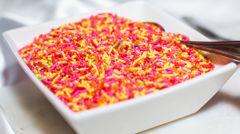 A square bowl of multicolored sprinkles with a spoon inside of it