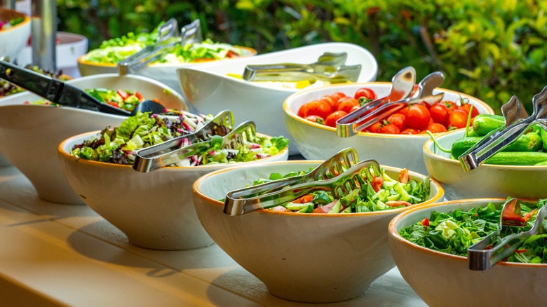 Several bowls of salads and additional garnish items on a buffet table with tongs.