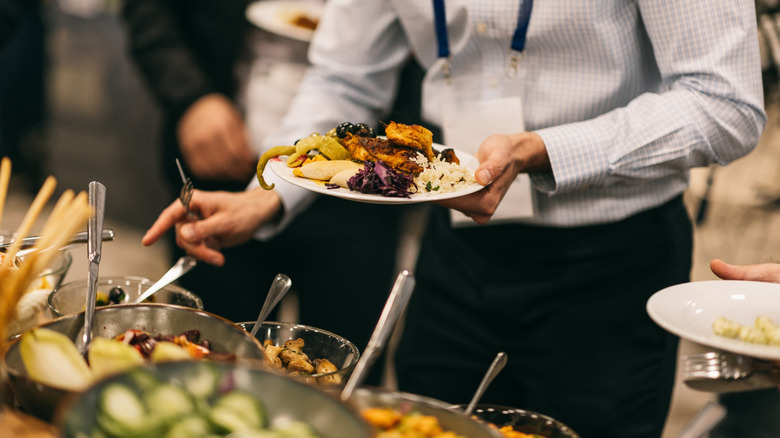 A man is standing at a buffet table holding a full plate, and reaching for a serving spoon.