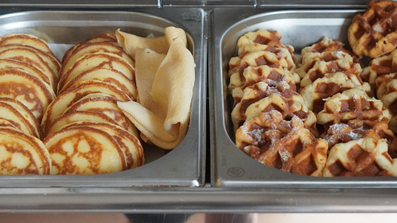A tray filled with pancakes, and a tray filled with waffles next to it set out on a buffet.