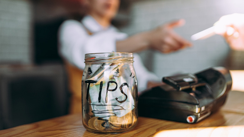 A glass jar labeled "TIPS" in front of a worker being handed something at the cash register