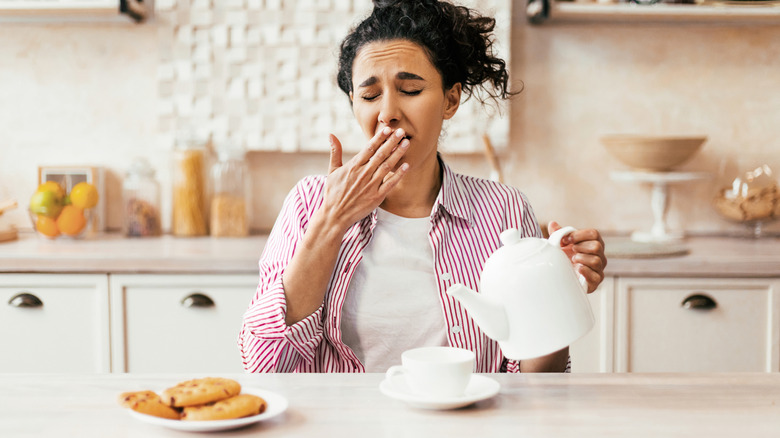 Person sitting in kitchen yawning as they pour from a white kettle into a white cup. Cookies on a plate are to the left.