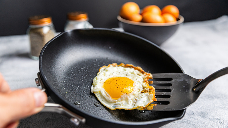 A cooked sunny-side-up egg with crispy edges being lifted with a spatula in a nonstick pan, with a bowl of eggs and salt and pepper shakers in the background and a bowl of brown eggs.