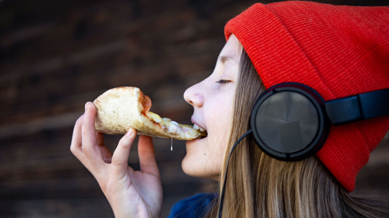 A young girl taking a bite of pizza while listening to music through her over-ear headphones