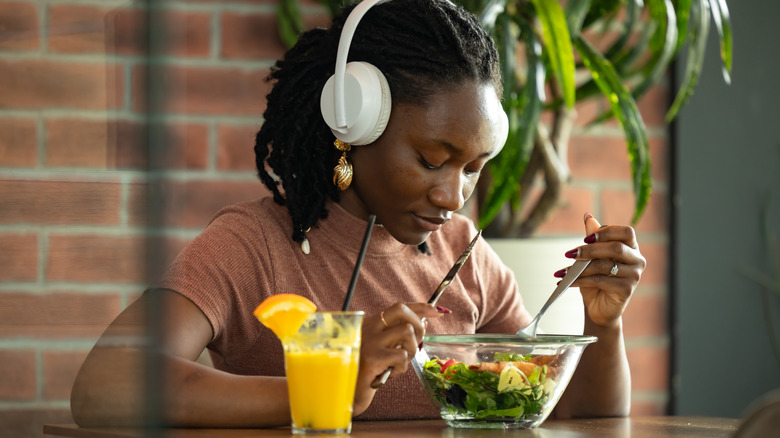 A young woman eating a salad with juice on the side and over-ear headphones on her ears