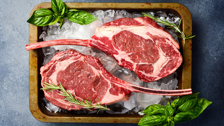 Two raw tomahawk steaks on ice with basil in a metal rectangular pan