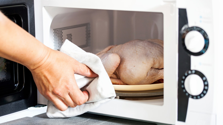 A hand uses a towel to grab a raw chicken inside a microwave