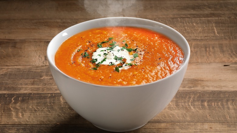 White bowl filled with tomato soup, garnished with fresh basil and cream with steam coming from it