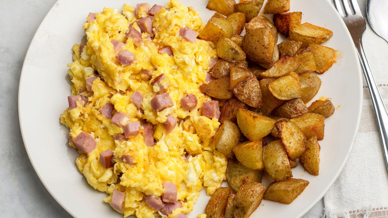 A white plate with scrambled eggs, pieces of diced ham, and golden brown potatoes on it.