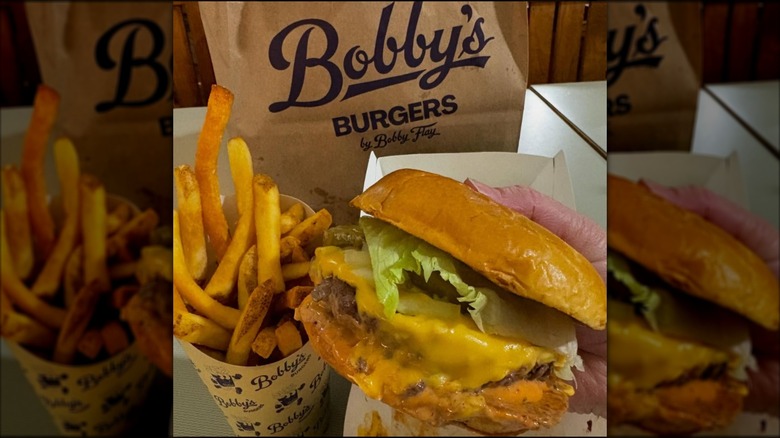 Someone holding a burger next to a cup of fries and a Bobby's Burgers-branded bag.