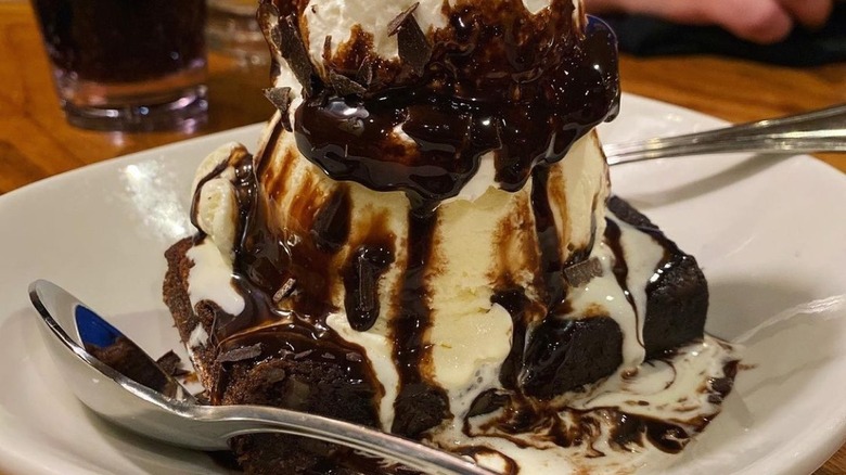 A plate with a slice of brownie topped with a scoop of vanilla ice cream, chocolate sauce and whipped cream with two spoons