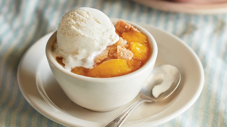Peach cobbler and ice cream in a small white bowl and plate with a spoon on the side