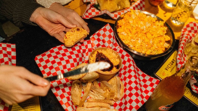 Hnads grabbing food from a busy table laden with popcorn, sandwiches, and pimento cheese