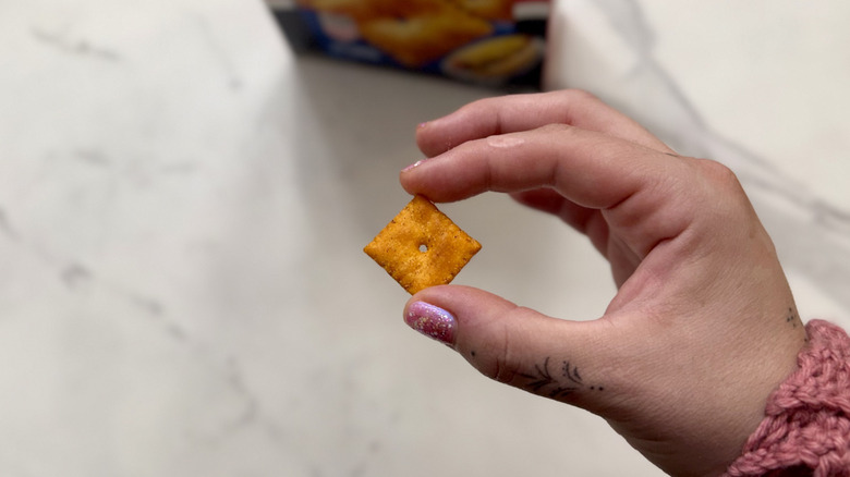 A hand holding a singular Cheez-It cracker above a countertop