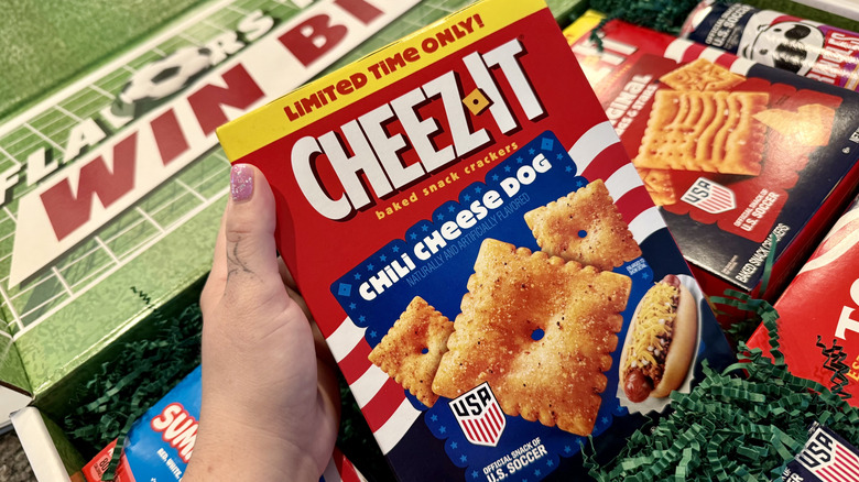 A hand holding a box of Cheez-Its in front of other snacks