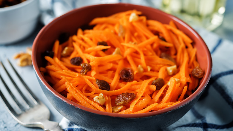 Shredded carrots and raisin salad with small walnut pieces in a brown bowl with the top of a fork next to it on a blue and white kitchen towel