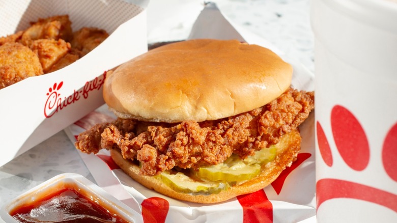 Chick-fil-A chicken sandwich on wrapper alongside a box of chicken nuggets, dipping sauces, and a drink