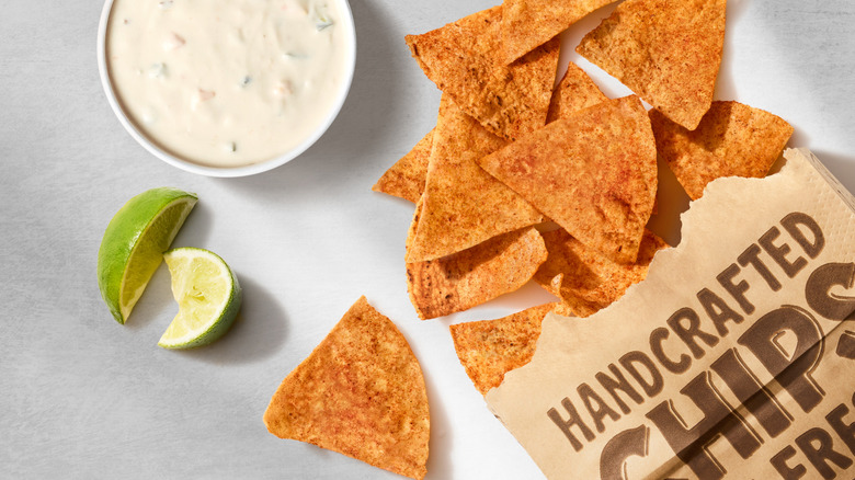 New Chipotle chili lime chips on surface next to white queso and lime wedges.
