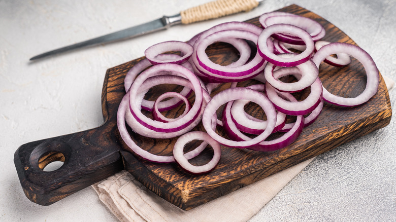 Sliced rings of red onion or purple onion on top of a wooden chopping board, with a knife and kitchen towel on either side.