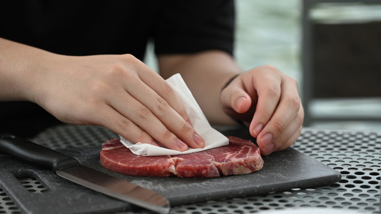 View of a person's hand as it blots a raw steak with a paper napkin or towel.