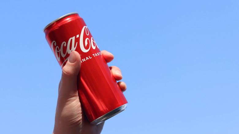 A hand holding up a can of Coca-Cola against a blue sky background.