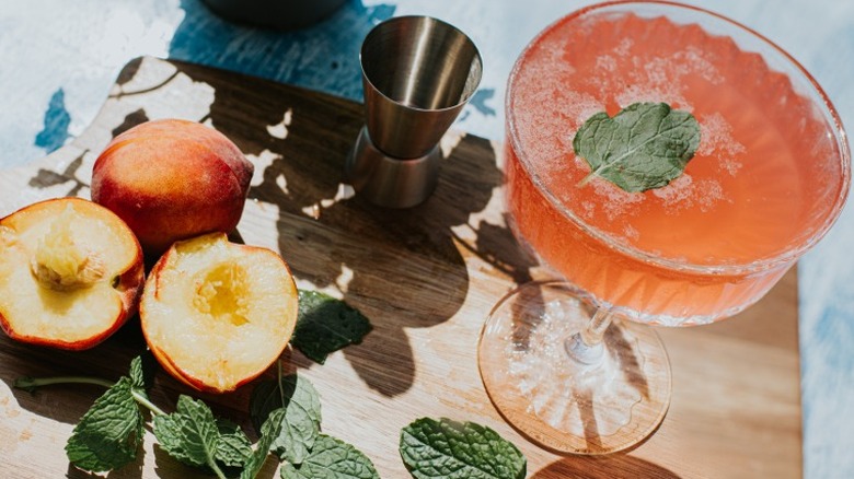 Seasonal cocktail in coupe glass with peaches and mint leaves on a cutting board.