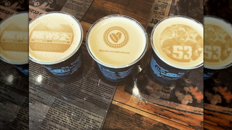 Three coffees in to-go cups on counter at Just Love Coffee shop, each with different latte art including an image of Keith Bulluck in 53 football jersey