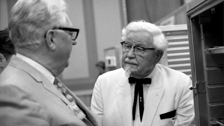 Colonel Sanders of KFC talking to a man in a suit in front of kitchen storage shelves.