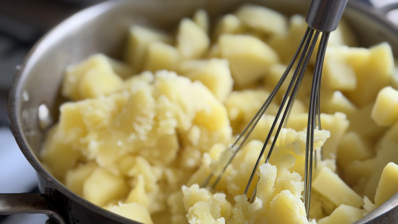 A wire whisk sits in a bowl of partially mashed potatoes