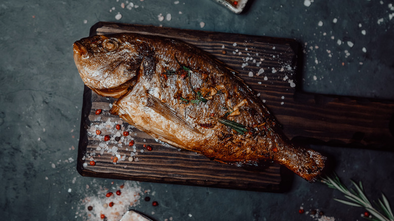 Whole grilled fish on a wooden board surrounded by salt and pepper