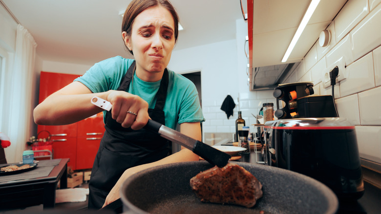 Woman with disappointed expression turning over overcooked tuna filet in pan on stovetop