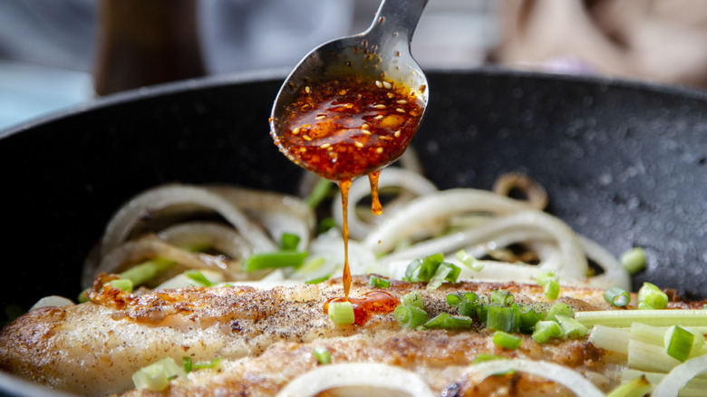 A cook spoons hot chile sauce over fish and vegetables in a frying pan