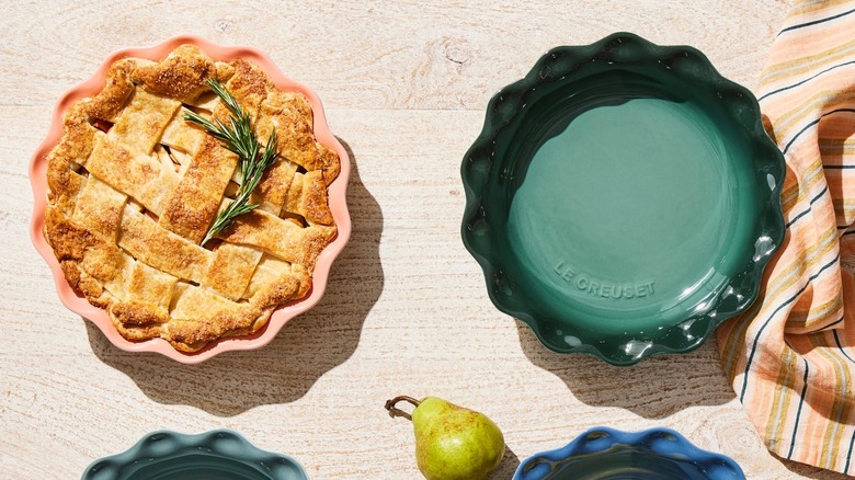 Empty green Le Creuset pie pan next to pink Le Creuset pie pan with latticed pie on wood surface