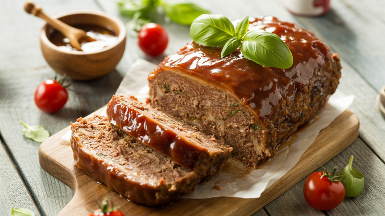 Homemade meatloaf with sauce and basil topping presented on a cutting board over wooden table.