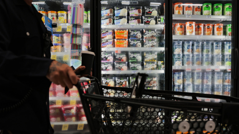 A supermarket shopper with cart passes by a refrigerated display holding different brands of ice cream