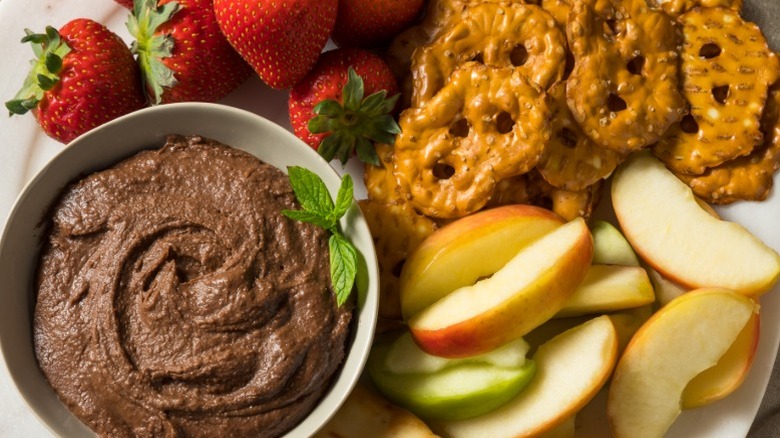 Bowl of homemade brownie batter hummus alongside strawberries, apple slices, and pretzel thins