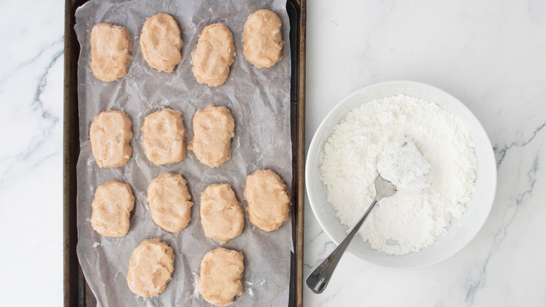 Raw chicken nuggets on baking sheet next to bowl of cornstarch with fork
