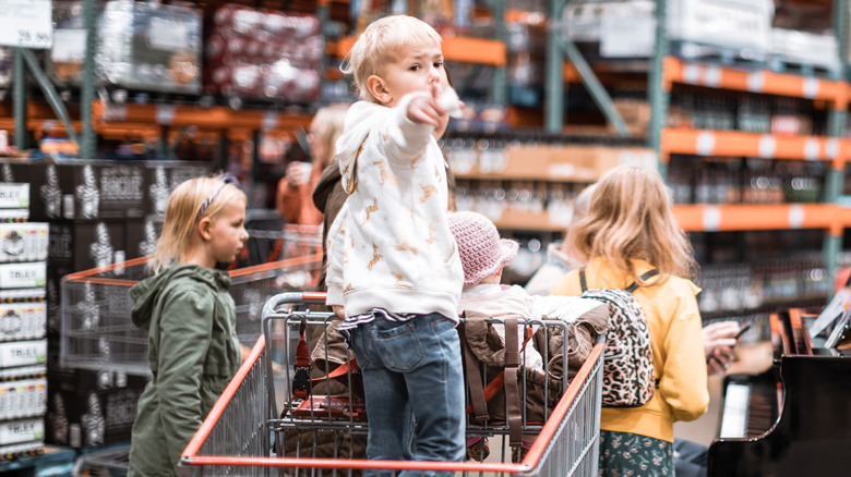 Boy standing in a Costco shopping cart and two girls standing next to it, with Costco shelves in the background