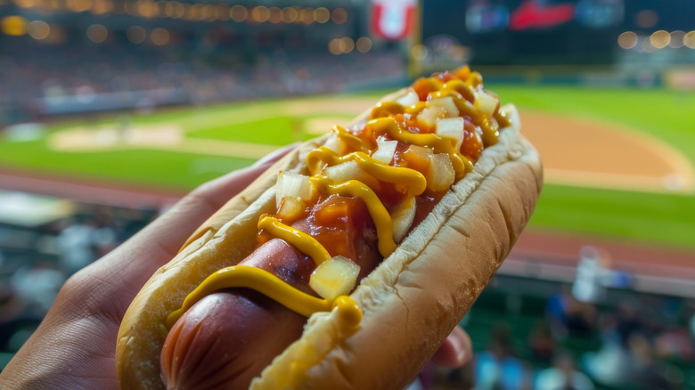 A hand holding a hot dog with a baseball stadium in the background