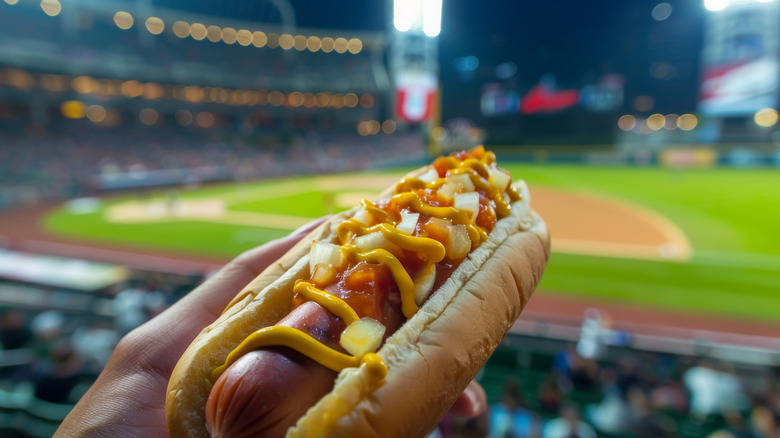 A hand holding a hot dog with a baseball stadium in the background