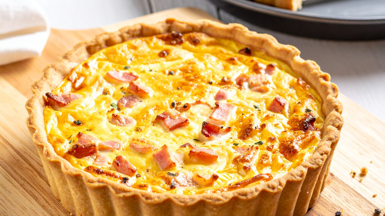 A whole quiche with ham bits sitting on a wood cutting board.