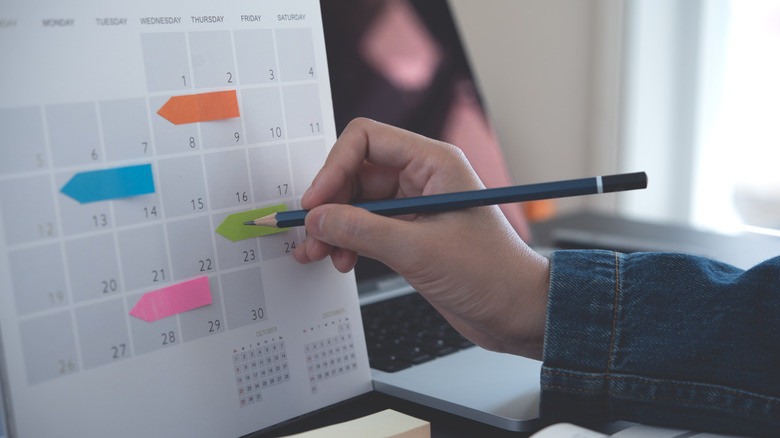 a hand holding a pencil moves to write something on a calendar page marked with colorful sticky notes
