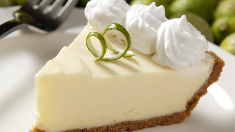 A closeup of a slice of key lime pie with limes in the background.