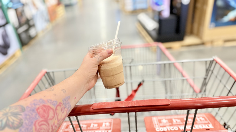 Hand holding a Costco Frappe in plastic takeout cup in front of Costco shopping cart in warehouse aisle